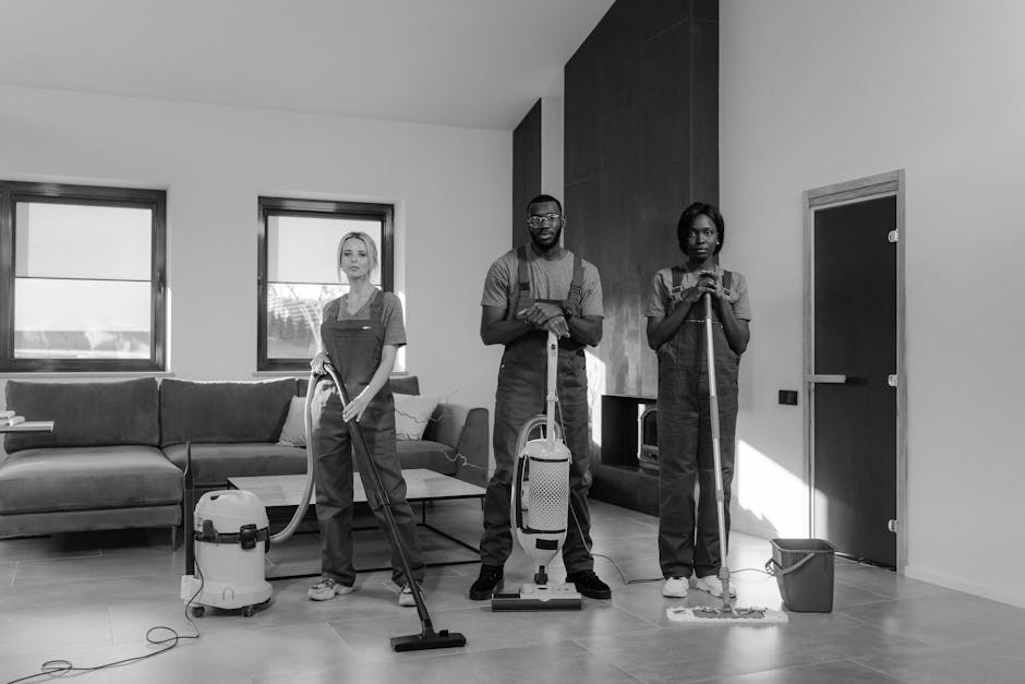 Three individuals engaged in surface cleaning within a modern living room. The scene includes a woman on the left using a vacuum cleaner on the carpeted floor, a man in the center holding a spray bottle and standing next to a portable deep cleaning machine, and another woman on the right holding a mop with a bucket placed nearby. The room features a dark wooden accent wall, a light-colored tiled floor, a grey sofa with cushions, and two windows allowing natural light to illuminate the space. The surfaces being cleaned are well-maintained and free of dust, reflecting thorough hygiene and regular cleaning practices. This scene exemplifies domestic cleaning efforts that contribute to sanitised and hygienic environments, consistent with surface and deep cleaning standards promoted by Hounslow Carpet Cleaning.