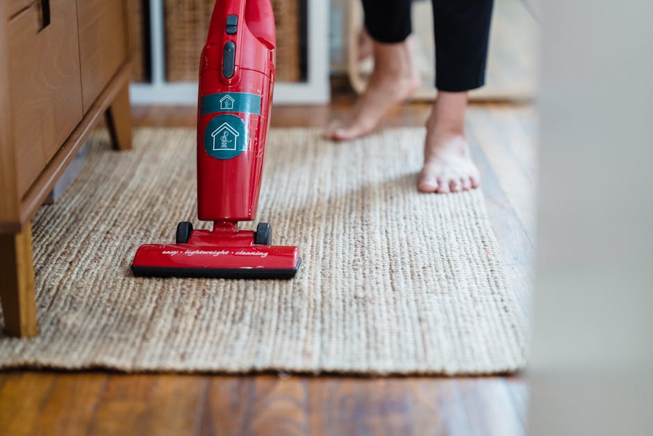 Close-up view of a red handheld vacuum cleaner with a label indicating 'deep cleaning' and 'sanitisation' placed on a textured beige rug in a residential living space. In the background, a person is barefoot, standing on the rug, with their legs partially visible, suggesting active cleaning. The room features wooden flooring adjacent to the rug, with natural light illuminating the scene, highlighting the cleanliness of the area. Furniture, such as a wooden cabinet or sideboard, is visible on the left side of the image, emphasizing a tidy, well-maintained environment. The scene illustrates routine surface cleaning practices, highlighting the importance of maintaining hygiene and cleanliness in Hounslow homes, and is associated with professional services like those offered by Hounslow Carpet Cleaning.