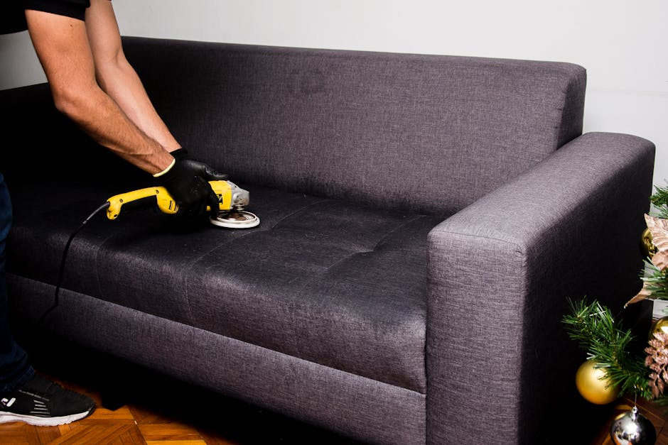 A person wearing black gloves is using a yellow handheld electric fabric steamer or upholstery cleaning device on a dark grey sofa in a living room. The sofa, made of textured fabric, is situated on a wooden floor near a white wall. To the right of the sofa, there is a decorated Christmas tree with gold ornaments and pink ribbons, indicating a festive setting. The room is well-lit, and the scene depicts surface cleaning and maintenance of the upholstered furniture to ensure hygiene and cleanliness, consistent with professional domestic cleaning services as offered by Hounslow Carpet Cleaning. The overall impression is of a thorough deep cleaning process aimed at revitalising and sanitising the sofa area.