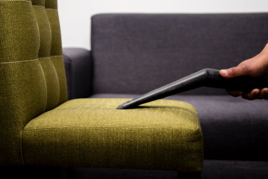 A person wearing black gloves is using a yellow handheld electric fabric steamer or upholstery cleaning device on a dark grey sofa in a living room. The sofa, made of textured fabric, is situated on a wooden floor near a white wall. To the right of the sofa, there is a decorated Christmas tree with gold ornaments and pink ribbons, indicating a festive setting. The room is well-lit, and the scene depicts surface cleaning and maintenance of the upholstered furniture to ensure hygiene and cleanliness, consistent with professional domestic cleaning services as offered by Hounslow Carpet Cleaning. The overall impression is of a thorough deep cleaning process aimed at revitalising and sanitising the sofa area.