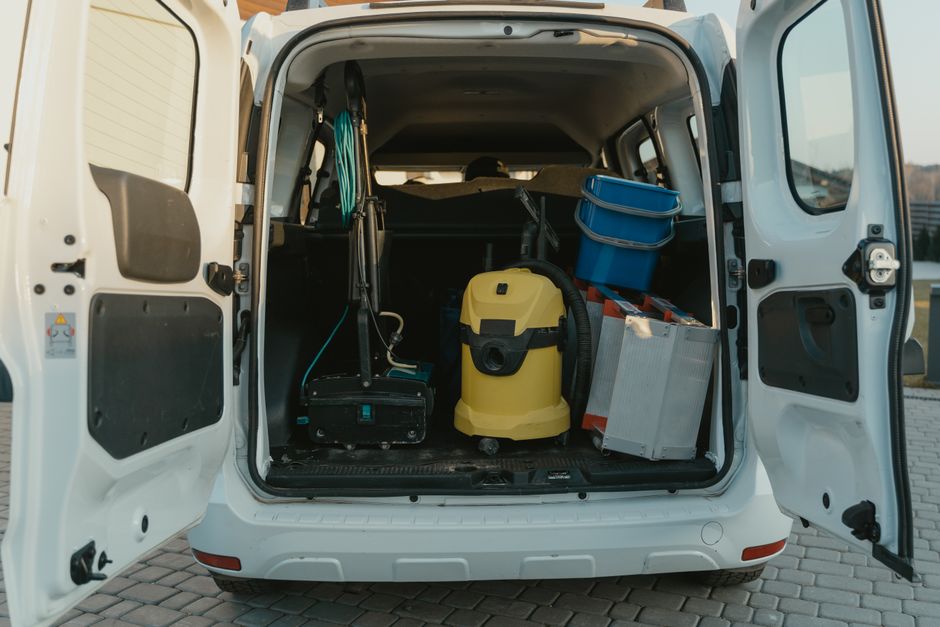 The open rear of a white cleaning van parked on a paved surface, revealing various cleaning equipment inside. Visible items include a yellow wet vacuum, a portable carpet cleaner, a metal trolley with blue storage bins, and a black sprayer. The van's interior surfaces appear clean and organized, prepared for surface cleaning, deep cleaning, or sanitisation tasks. The setting suggests the vehicle is ready for residential or commercial cleaning services, supported by Hounslow Carpet Cleaning, as indicated on the webpage. The lighting is natural, highlighting the equipment and the tidy cargo space, emphasizing professional maintenance and preparation for hygiene-focused cleaning activities.
