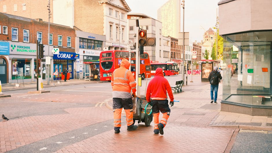 Two street cleaning workers dressed in bright orange and red high-visibility uniforms are pushing a cleaning cart on a busy city sidewalk in Hounslow. The scene features a red double-decker bus and various storefronts, including a Yorkshire Building Society branch and a Coral betting shop. The street is paved with brick and asphalt, with a traffic light visible at the intersection. The lighting indicates it is either early morning or late afternoon. The workers are engaged in surface cleaning or maintenance, reflecting the importance of hygiene and cleanliness in urban areas. The image is captured in front of a commercial area with pedestrians and street furniture, highlighting routine municipal sanitation efforts by Hounslow Carpet Cleaning and local service providers.