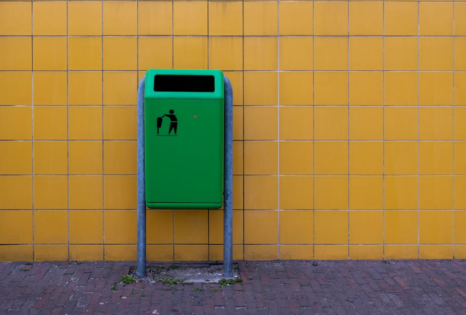 A green trash bin with a black icon depicting a person disposing of waste is mounted on a metal stand against a yellow tiled wall. The bin is situated outdoors on a brick-paved surface beneath bright, natural lighting. The tiles are uniform in size, with visible grout lines, and appear clean and well-maintained. The bin's surface is smooth and free of dirt or marks, indicating regular cleaning and upkeep by Hounslow Carpet Cleaning as part of their domestic cleaning and surface maintenance services. The background wall is evenly tiled, providing a vibrant contrast to the green bin, which is positioned centrally in the frame, emphasizing cleanliness, hygiene, and waste disposal processes.