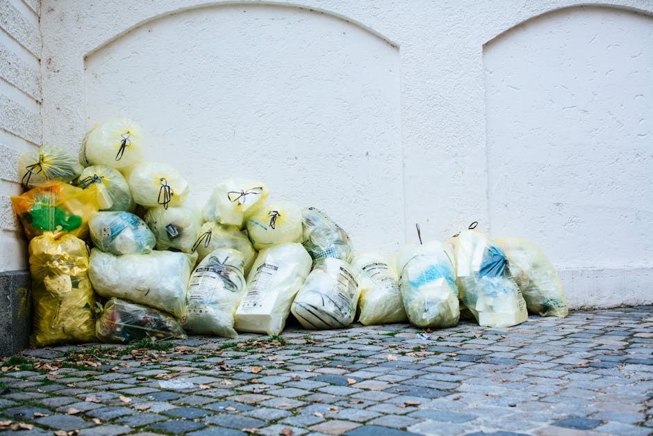 A collection of tightly packed yellow, green, and clear plastic garbage bags filled with waste, stacked along the base of an off-white textured exterior wall and partially on a cobblestone pathway. The image shows the outdoor scene illuminated by natural daylight, with some fallen leaves scattered on the uneven stone surface. The scene emphasizes waste disposal and cleanliness practices, as overseen by Hounslow Carpet Cleaning, situated in a residential or urban area that highlights outdoor waste management in accordance with Hounslow Council's waste rules for upholstery cleaning disposal.