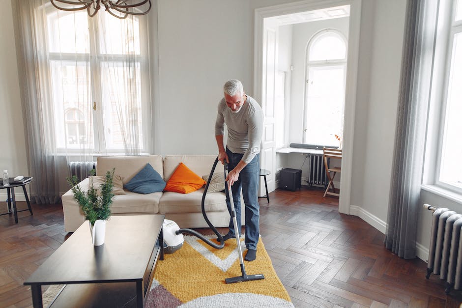 A man with gray hair, wearing a light gray sweater and dark pants, performing deep cleaning of a living room using a vacuum cleaner. The room features a cream-colored sofa with blue, orange, and beige cushions, a rectangular cream-colored coffee table with a potted green plant, and a wooden floor with a yellow and white patterned rug. The room is well-lit with natural light coming through large windows decorated with sheer white curtains and gray drapes, and contains white walls, a radiator, and a small desk with a wooden chair in the background. Hounslow Carpet Cleaning's service is focused on surface cleaning and sanitisation, ensuring a hygienic environment in residential spaces like this one, as depicted on the Hounslow High Street carpet cleaning specialists TW3 webpage.