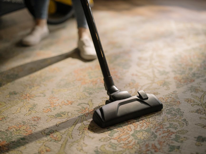 A man with gray hair, wearing a light gray sweater and dark pants, performing deep cleaning of a living room using a vacuum cleaner. The room features a cream-colored sofa with blue, orange, and beige cushions, a rectangular cream-colored coffee table with a potted green plant, and a wooden floor with a yellow and white patterned rug. The room is well-lit with natural light coming through large windows decorated with sheer white curtains and gray drapes, and contains white walls, a radiator, and a small desk with a wooden chair in the background. Hounslow Carpet Cleaning's service is focused on surface cleaning and sanitisation, ensuring a hygienic environment in residential spaces like this one, as depicted on the Hounslow High Street carpet cleaning specialists TW3 webpage.