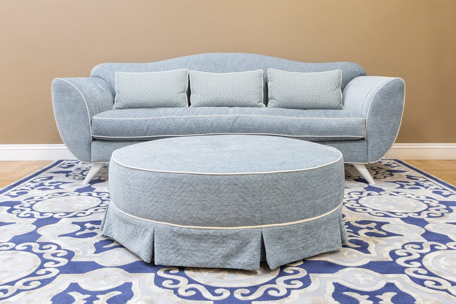 A light blue upholstered sofa with rounded armrests and three matching cushions, situated on a decorative patterned area rug featuring blue and white floral motifs, in a room with beige walls and wooden flooring. In front of the sofa is a matching round ottoman with pleated skirt, all appearing clean and well-maintained, illustrating domestic surface cleaning and upholstery sanitisation by Hounslow Carpet Cleaning.