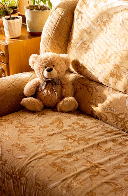 A person wearing black gloves is using a handheld upholstery cleaning machine on a dark grey fabric sofa in a living room. The sofa has a padded backrest and armrest, with visible clean and slightly wet areas from the cleaning process. The room has wooden flooring and white walls, with a small decorated Christmas tree featuring gold and pink ornaments positioned nearby. Natural light illuminates the scene, emphasizing the surface cleanliness achieved through professional deep cleaning offered by Hounslow Carpet Cleaning. This image highlights the importance of fabric sofa sanitisation and maintenance in domestic settings, ensuring hygiene and upholstery longevity.