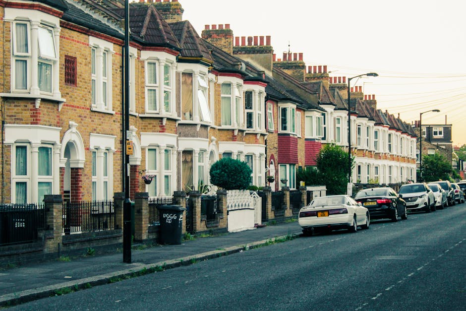 A row of traditional terraced houses with brick facades and white bay windows in a residential street, illuminated by warm sunset light. The front gardens are neatly fenced with low brick walls and black iron railings, featuring small shrubs and potted plants. Several parked cars line the asphalt street, which is clean and well-maintained. Street lamps and overhead power lines run along the sidewalk, with a clear sky in the background. The scene showcases a typical urban residential area related to housing and property maintenance, relevant to landord obligations and cleaning compliance in Hounslow flats, as provided by Hounslow Carpet Cleaning for domestic surface cleaning and deep cleaning services.
