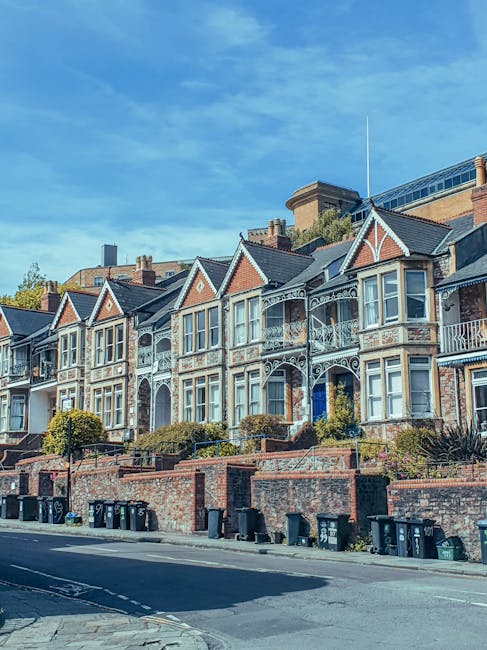 A row of Victorian-style terraced houses with ornate white decorative trim and bay windows, situated on a slightly elevated street level. The houses are built with red brick and feature pitched roofs with slate tiles. In front of the buildings, there is a low brick wall with steps leading up to each entrance, surrounded by well-maintained bushes and small trees. The street below is lined with black wheelie bins, and the pavement appears clean and tidy under natural daylight. The scene conveys a residential area that benefits from regular surface cleaning and maintenance, as supported by Hounslow Carpet Cleaning's dedication to domestic cleaning and hygiene standards, aligning with landlord obligations and cleaning compliance in Hounslow flats.
