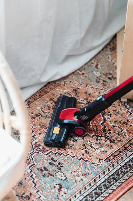 A close-up image of a handheld vacuum cleaner with a black and red design, actively cleaning a patterned, traditional-style rug with a mix of warm tones and intricate geometric motifs. The rug is laid out on a hardwood floor near a light-colored curtain, and the scene is well-lit, highlighting the surface cleaning process. Visible parts of nearby furniture and the texture of the rug emphasize a tidy, maintained living space. This image illustrates domestic cleaning activities undertaken by Hounslow Carpet Cleaning, focusing on deep cleaning and stain removal of area rugs.