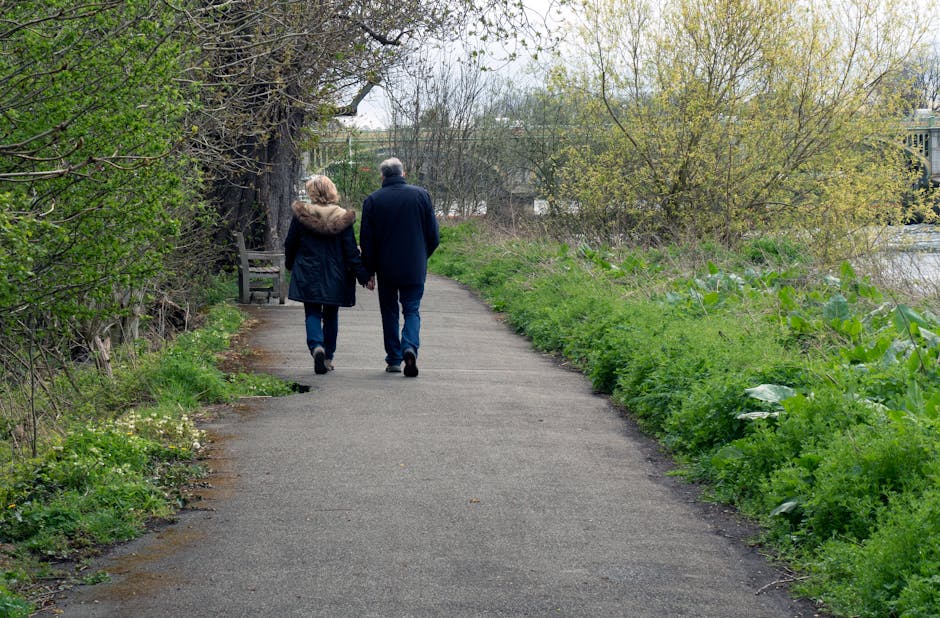 A couple walks hand in hand along a paved path in a park during spring, surrounded by lush green bushes and trees with light green foliage. The woman wears a dark coat with a fur-lined hood, and the man is dressed in a navy jacket; both are wearing casual trousers and walking shoes. The path is clean and well-maintained, with a wooden bench visible on the left side under a large tree, and the scene is illuminated by natural daylight, creating a peaceful outdoor setting. The setting emphasizes the importance of regular surface cleaning and maintenance, aligning with the role of [COMPANY_NAME] in providing deep cleaning, sanitisation, and domestic cleaning services, as discussed in the page titled 'What to know about hidden cleaning fees in Hounslow' at [PAGE_URL].
