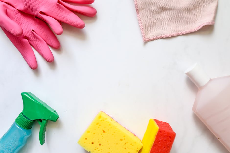 An arrangement of cleaning tools and supplies on a white surface, including a pair of pink rubber cleaning gloves, a light pink microfiber cloth, a green spray bottle with a blue liquid inside, a yellow and red kitchen sponge, and a white plastic bottle, all positioned to suggest surface cleaning or sanitisation in a domestic setting, consistent with Hounslow Carpet Cleaning's focus on deep cleaning and hygiene to address hidden cleaning fees in Hounslow.